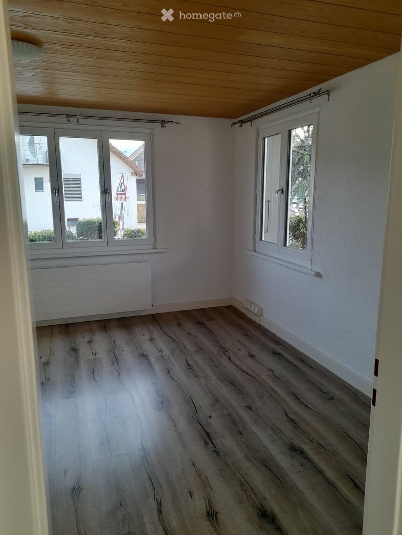 Empty room with light wooden flooring, white walls, large windows, and a radiator. The ceiling has a wooden panel design.