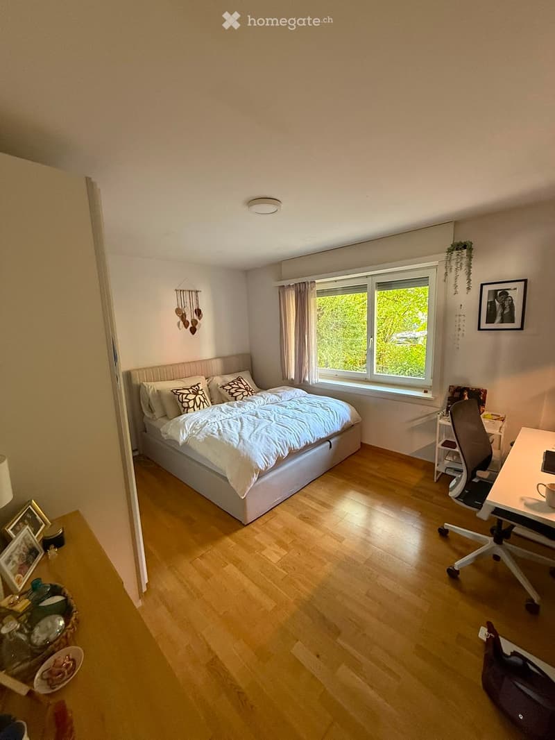 Cozy bedroom with a large bed, wooden floor, desk, chair, and decorative items including picture frames and a wicker basket.
