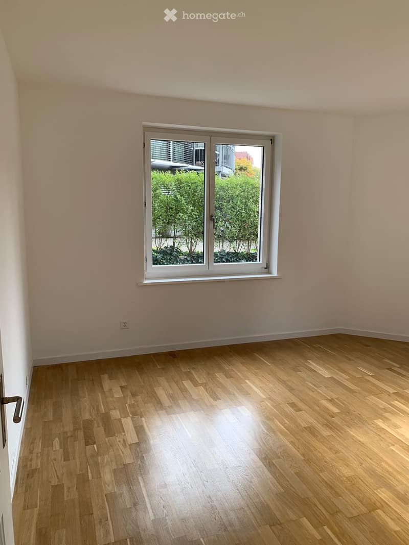 Empty room with light wooden flooring and a large window with white frame, showing greenery outside.