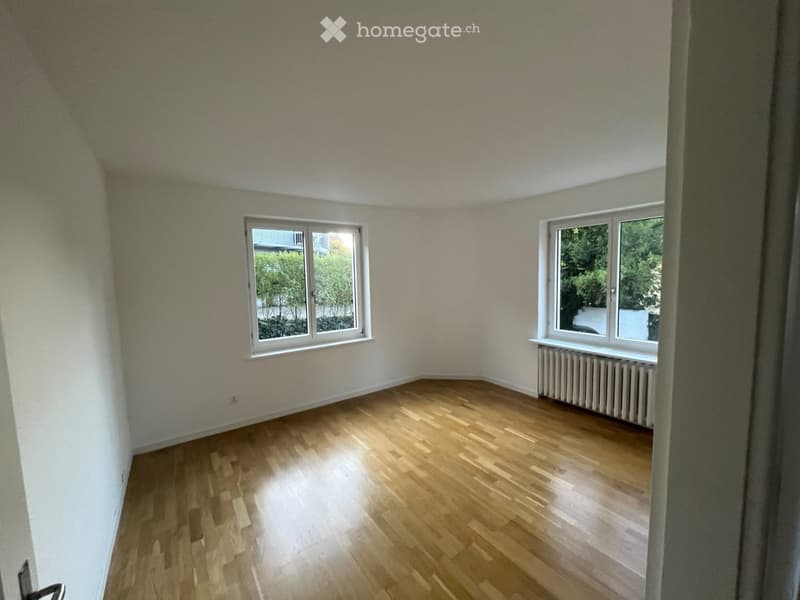 Empty room with wooden flooring, white walls, and two windows offering a view of greenery outside.