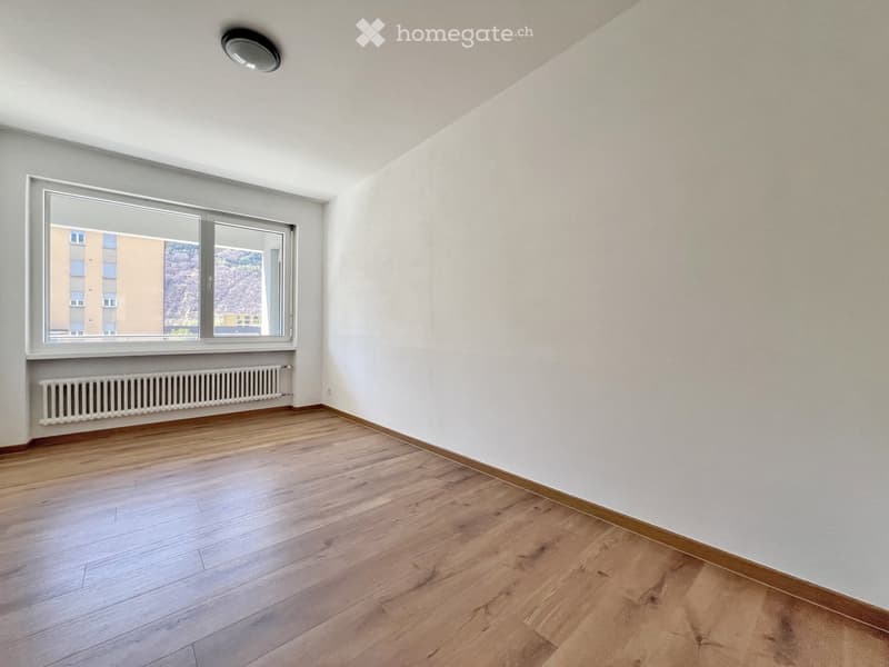 Empty room with wooden flooring, white walls, a large window with a view, and a radiator below the window.