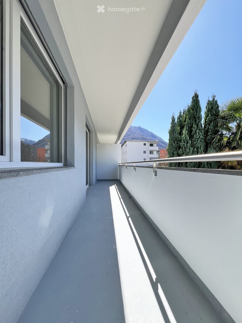 Modern balcony with white walls, grey floor, metal railing, and large windows overlooking a scenic mountain view.
