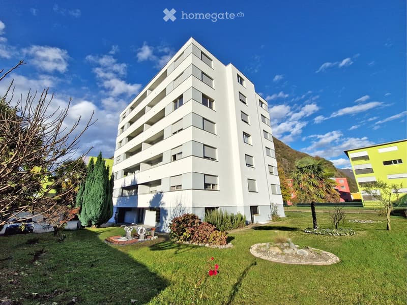 Modern multi-story apartment building with white facade, balconies, and surrounding greenery including trees and shrubs.