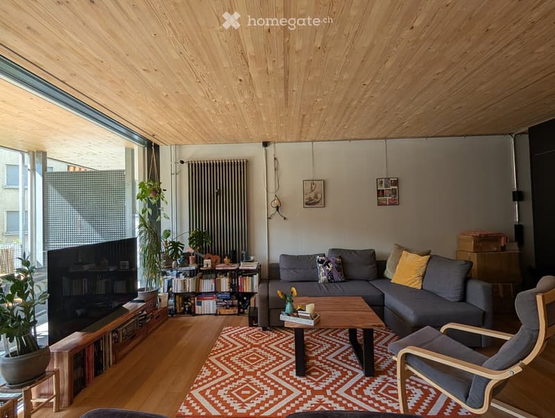 Modern living room with wooden ceiling, large window, grey sectional sofa, wooden coffee table, armchair, bookshelf, and plants.