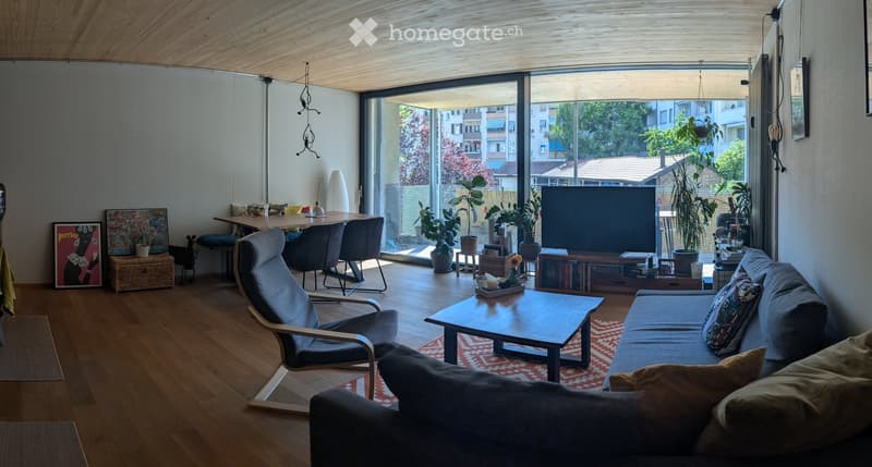 Modern living room with wooden floors, large windows, a grey sofa, coffee table, chairs, dining table, TV, and several potted plants.
