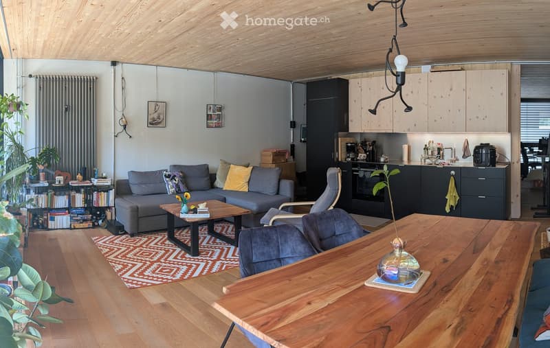 Open-plan living and dining area with wooden floors, grey sofa, coffee table, dining table, chairs, bookshelf, kitchen area with cabinets, oven, and fridge.