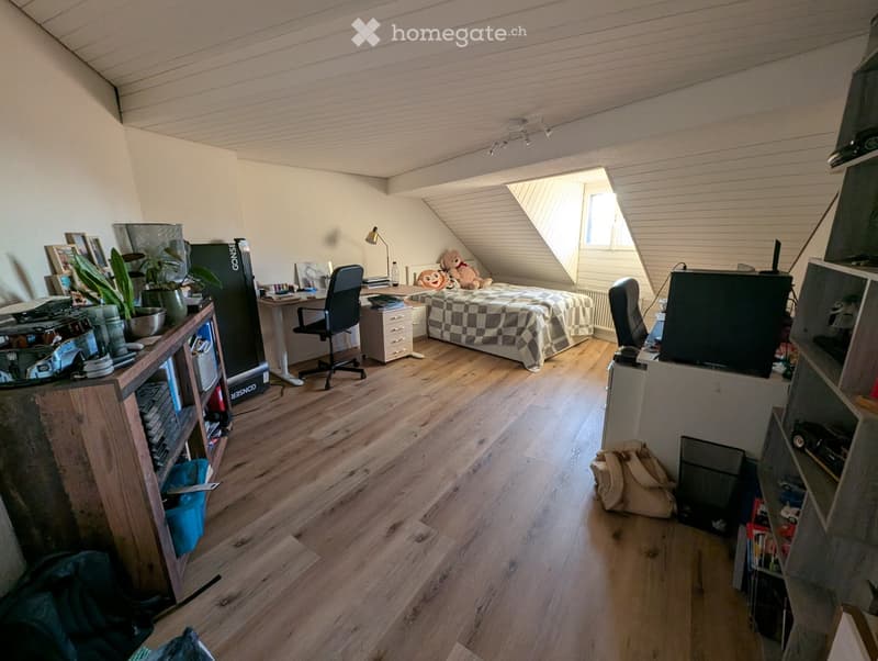 A cozy attic bedroom with wooden flooring, a slanted ceiling, and a large window. Features a bed with a checkered blanket, a desk with a chair, a TV, and shelves with books and decorative items.