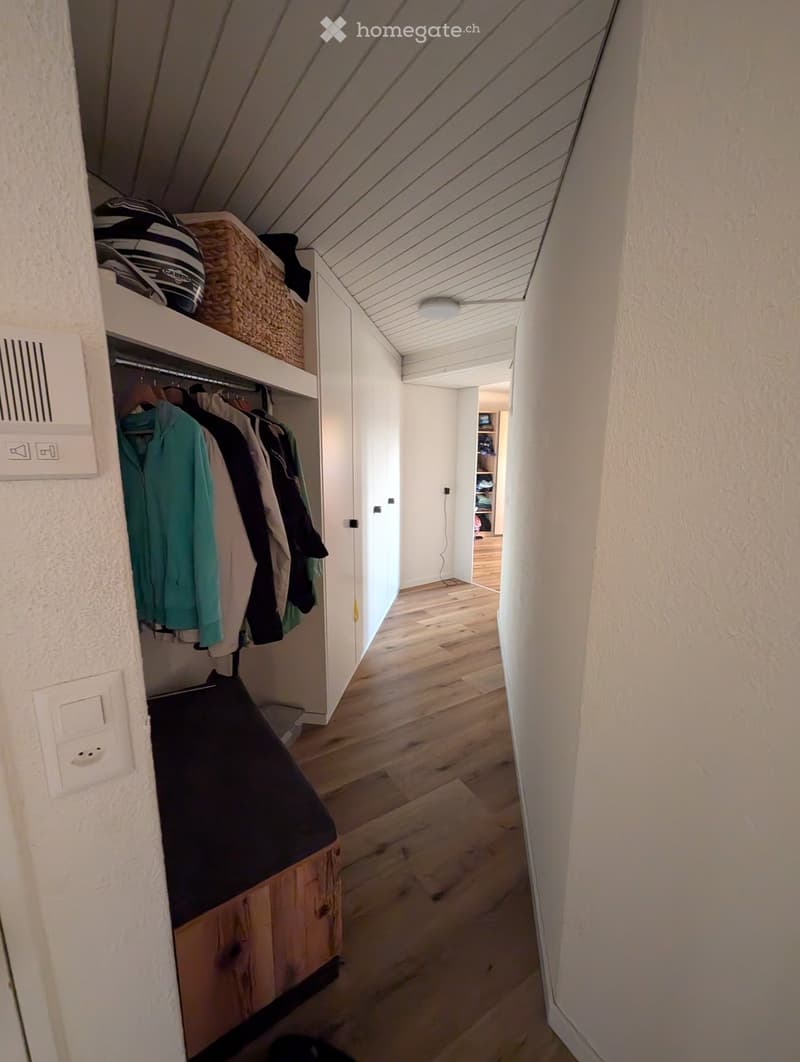 A narrow hallway with wooden flooring, white walls, and built-in storage including a closet with hanging clothes and a shelf with baskets. The ceiling has exposed wooden beams and a recessed light fixture.