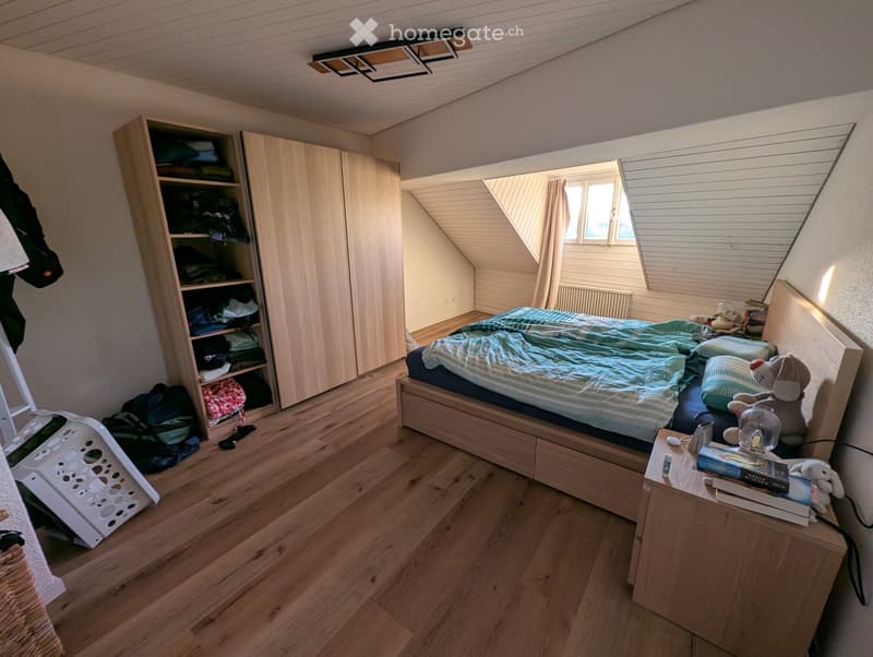 Cozy attic bedroom with wooden floors, slanted ceilings, and large windows. Features a double bed with blue bedding, a nightstand with books and a toy, a wardrobe, and a storage basket on the floor.