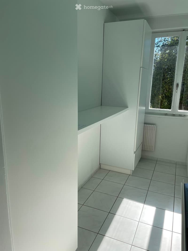 Empty room with white tiled flooring, white walls, and a built-in cabinet. A window with a view of greenery is visible, along with a radiator below it.