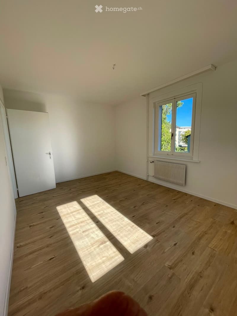 Empty room with wooden flooring, white walls, a window with a view of greenery, and a radiator below the window. The room has a door on the left side.