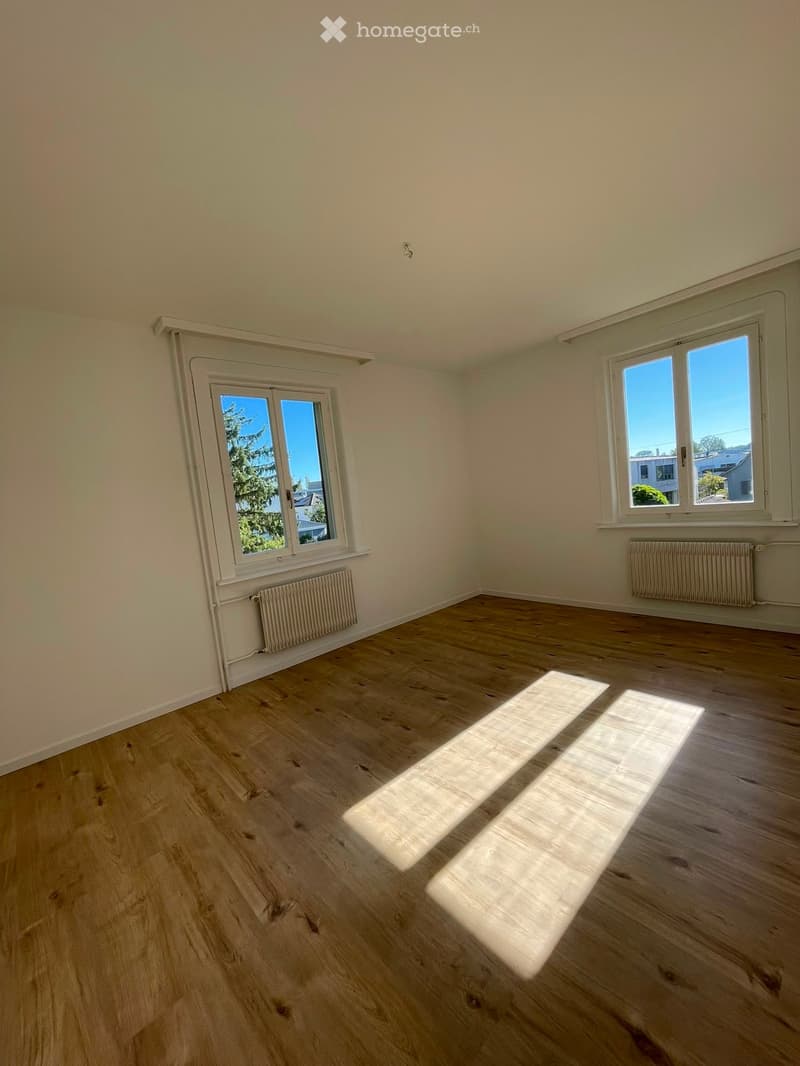 Empty room with wooden flooring, white walls, and two windows letting in natural light. Radiators are visible under the windows.