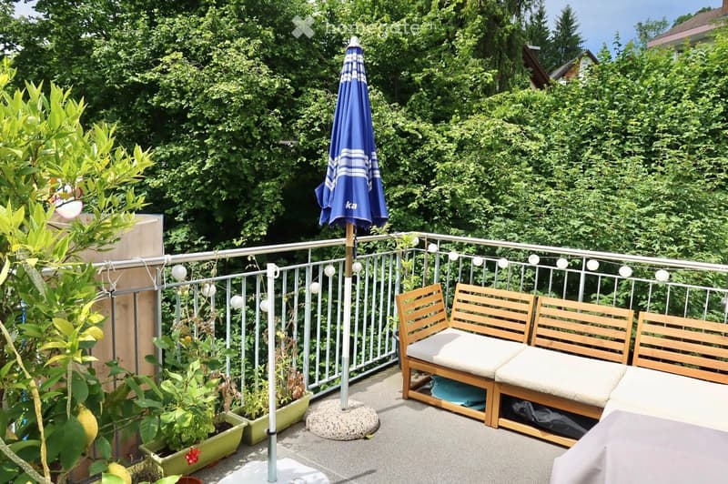 Balcony with wooden bench seating, umbrella, string lights, and potted plants.