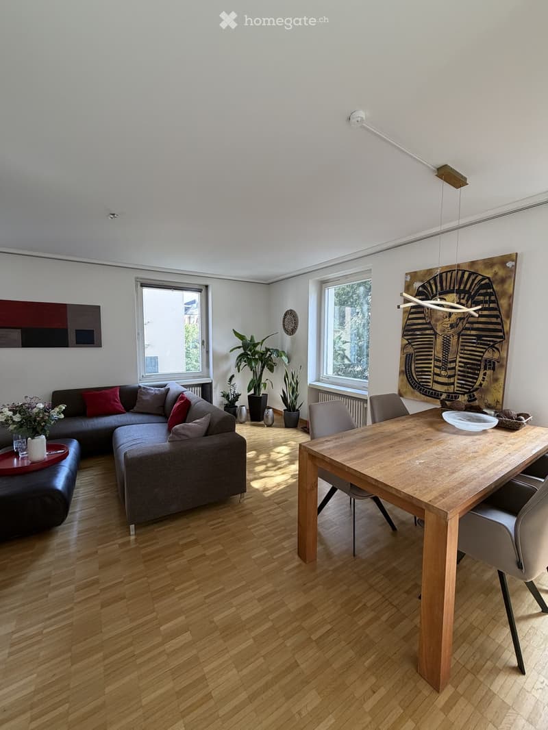 Modern living and dining area with a grey sectional sofa, wooden dining table, chairs, and decorative plants. The space features large windows, a wooden floor, and contemporary artwork on the walls.