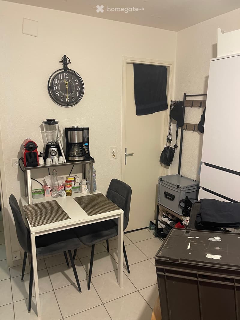 A small, furnished room with a table, two chairs, a refrigerator, a coffee maker, a blender, and a clock on the wall.