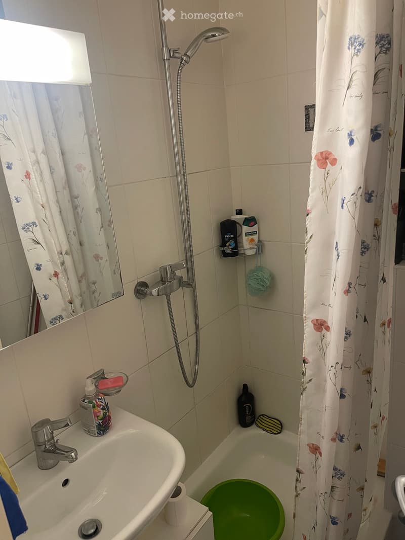 Bathroom with a bathtub, showerhead, sink, and various toiletries including a bottle, cup, and soap dish. The shower curtain is floral-patterned, and a mirror is mounted on the wall.