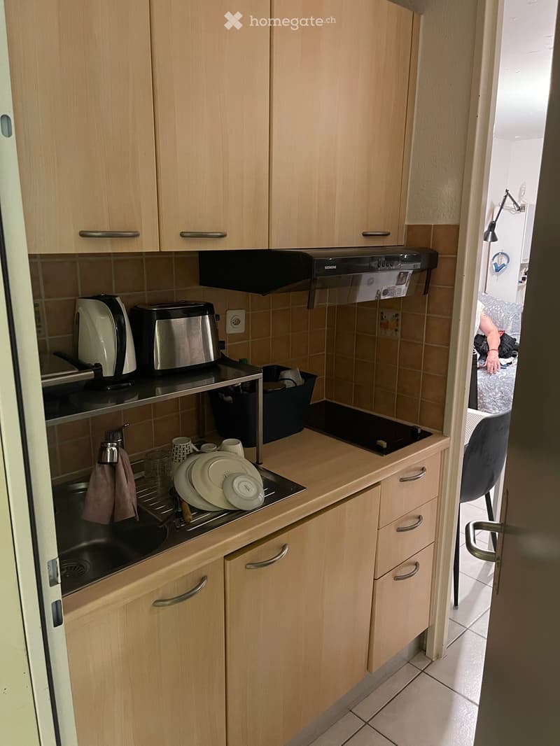 Compact kitchen with light wood cabinets, stainless steel appliances, and a small sink area with dishes and a dish rack.