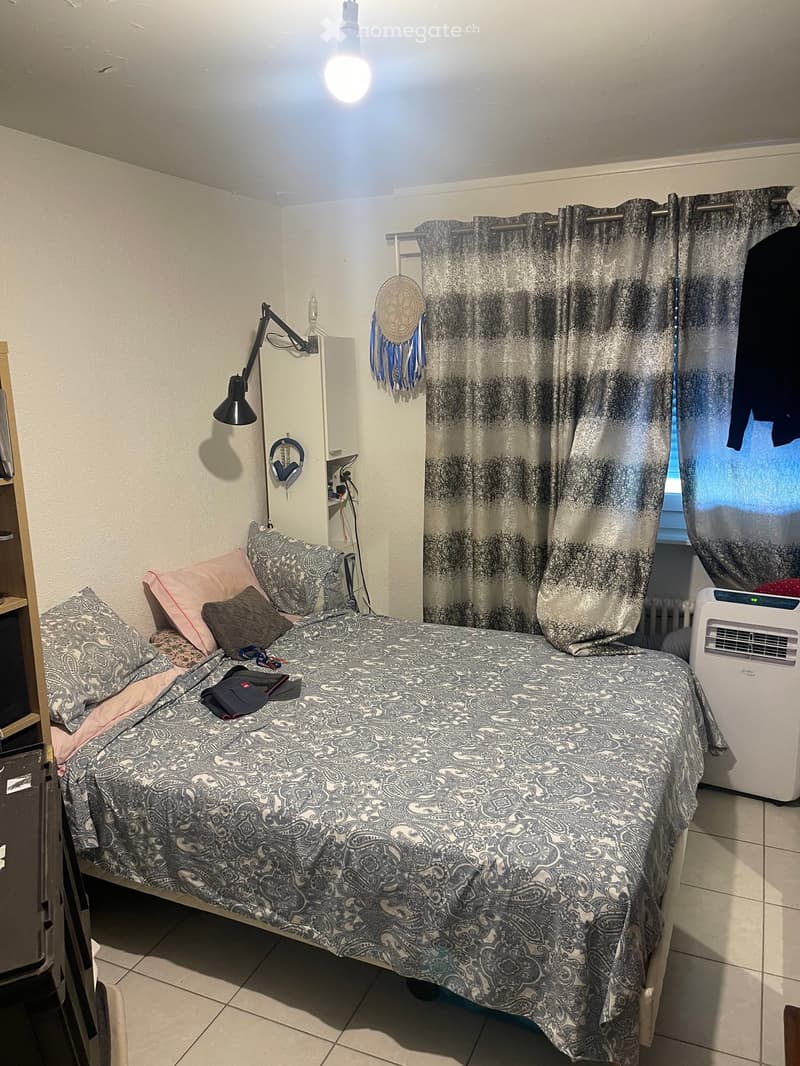 A small bedroom with a single bed, patterned bedding, a window with curtains, a wall-mounted lamp, a radiator, and an air conditioner unit.