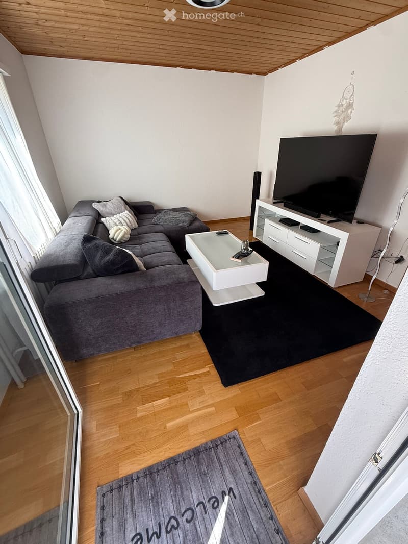Modern living room with a grey sectional sofa, white coffee table, TV stand with a large flat-screen TV, and wooden flooring.