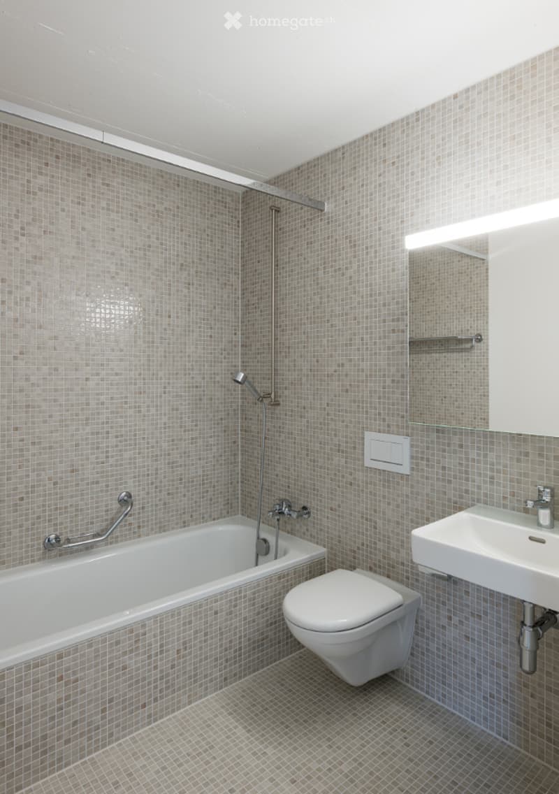 Modern bathroom with beige tiled walls and floor, featuring a bathtub with a handheld shower, a toilet, a sink with a mirror, and a built-in shelf above the sink.