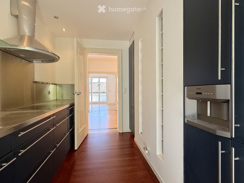 modern kitchen with wooden floors, stainless steel appliances, dark cabinets, bright lighting, glass extractor hood, and a large doorway leading to another room