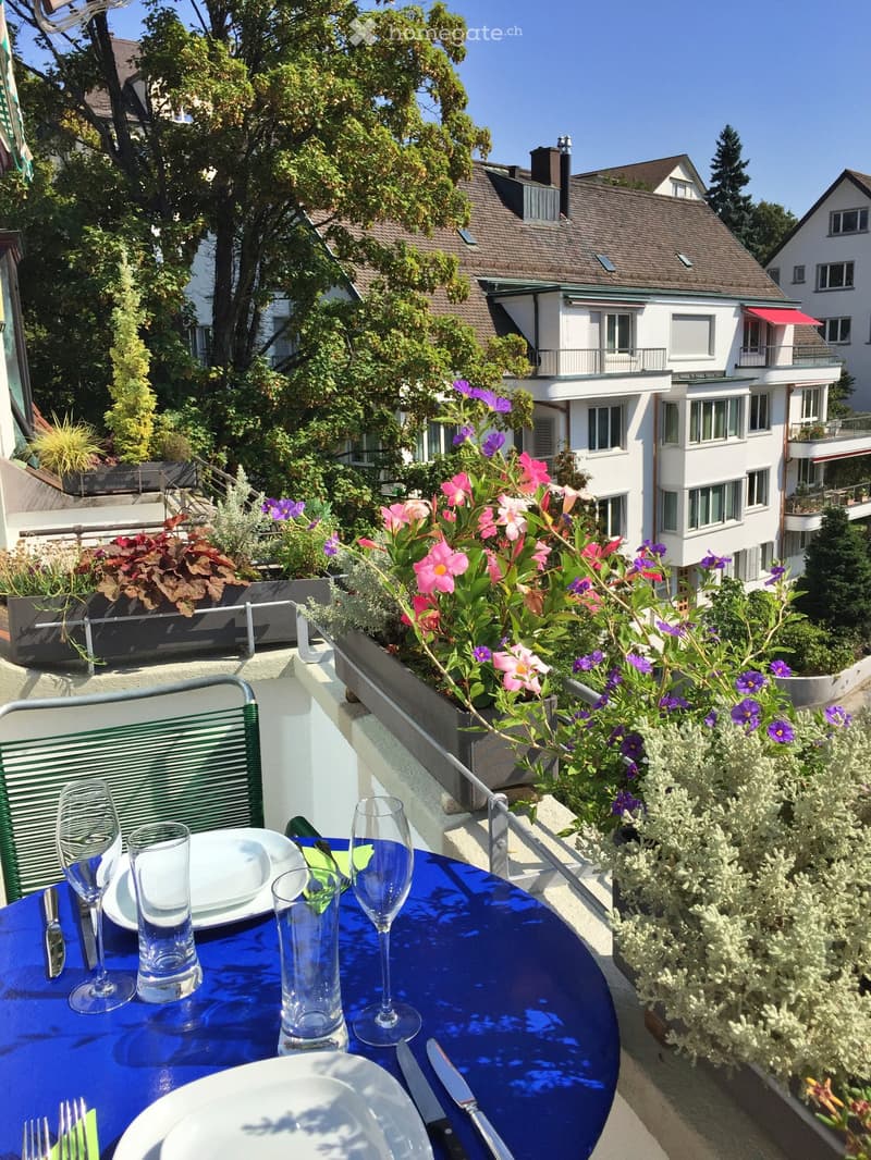 Outdoor dining area on a balcony with a blue table, plants, and a view of a building with a terrace and plants.