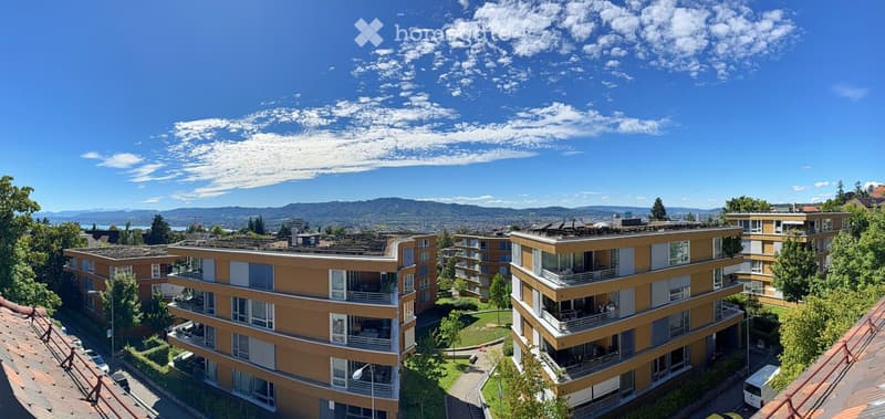 Aerial view of apartment buildings in a mountainous region with a clear blue sky and distant mountains, situated among greenery