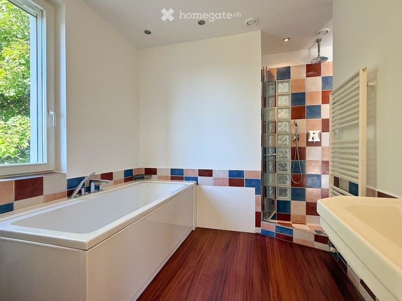large bathtub, white sink, colorful tiled wall, shower enclosure, shower head, towel rack, open window with view to trees
