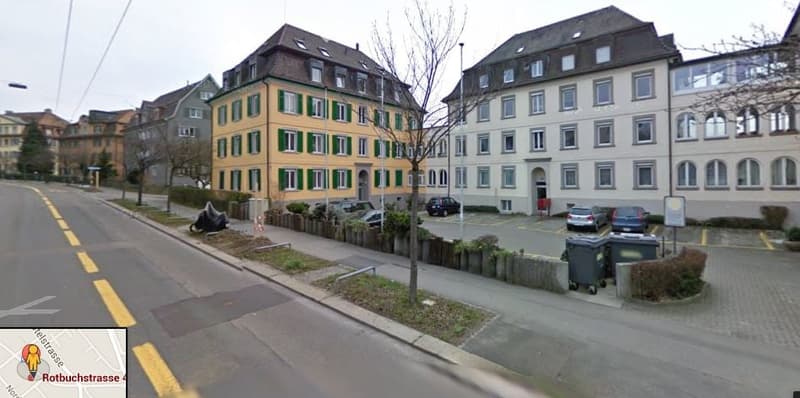 Multi-story residential building with green shutters and white trim, located on a street with parked cars and a small garden area.