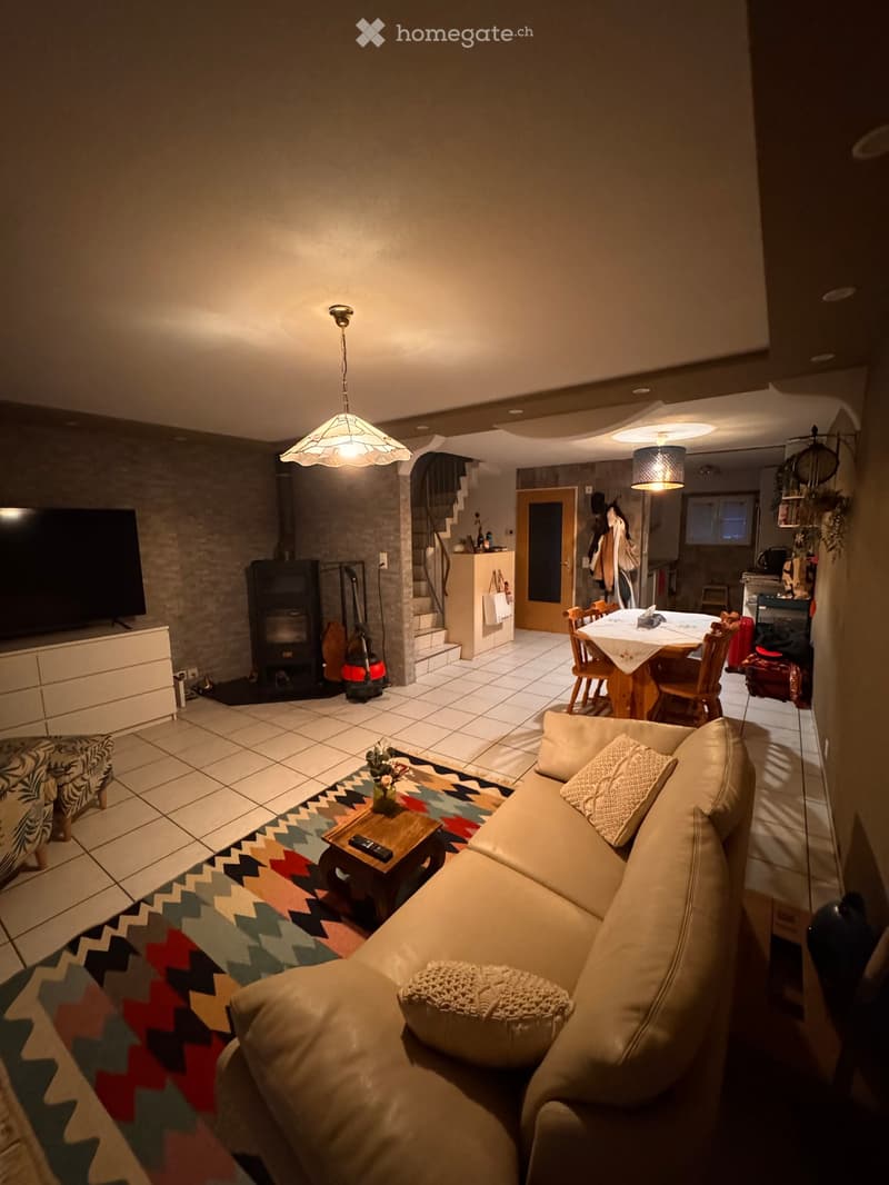 Living room with a beige sofa, coffee table, colorful rug, fireplace, TV, and dining area visible in the background.