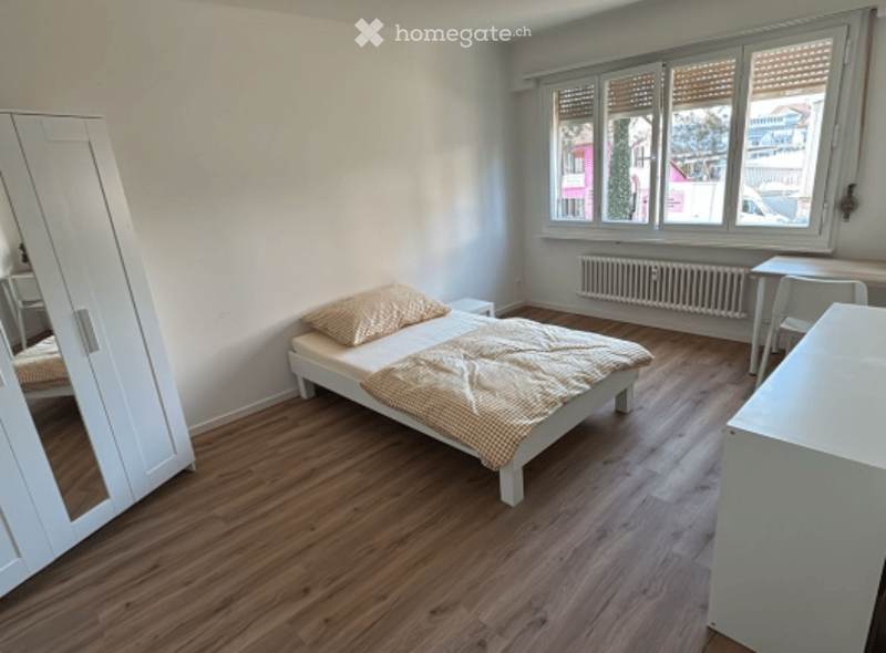 Cozy bedroom with wooden flooring, a single bed with a patterned blanket, a white wardrobe with a mirror, and a small table near the window.