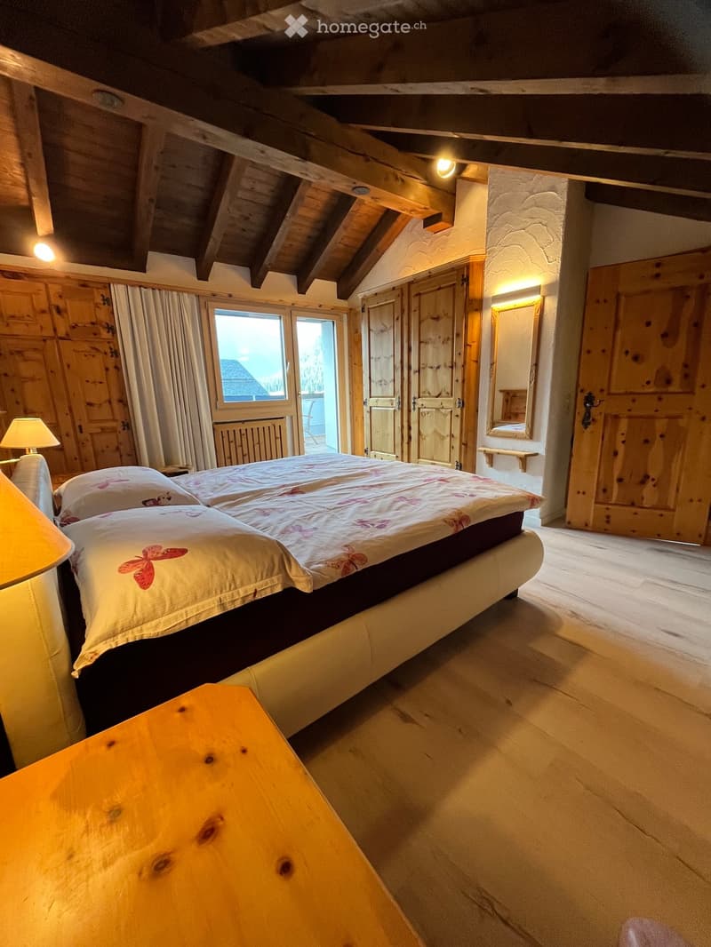 Cozy bedroom with wooden beams, a large bed with floral bedding, a wooden table, and warm lighting.