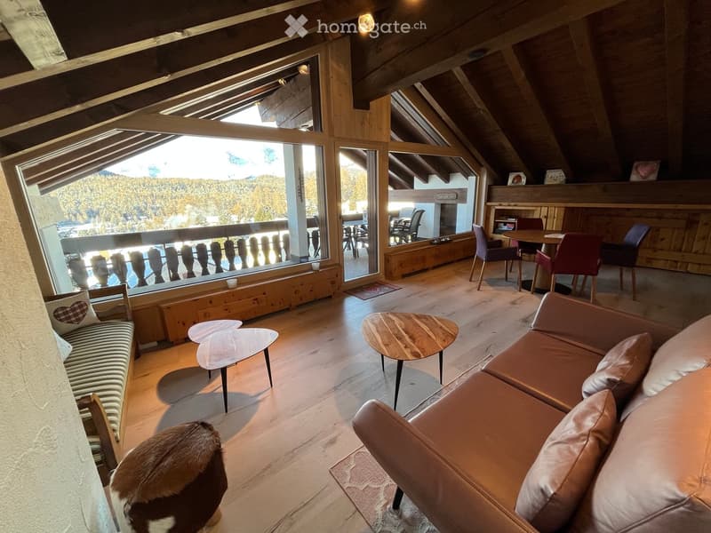 A spacious, well-lit living room with wooden floors, large windows offering a scenic mountain view, a leather sofa, a coffee table, a bench, and a dining area with a table and chairs.