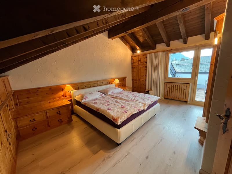 Cozy attic bedroom with wooden beams, a large bed with floral bedding, nightstands with lamps, and a wooden wardrobe. The room features exposed wooden beams and a large window with a view.