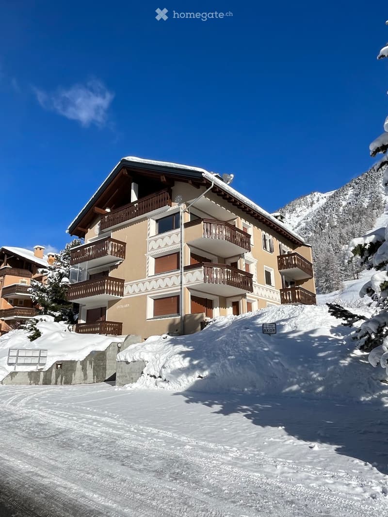 A multi-story residential building with balconies, surrounded by snow-covered trees and mountains under a clear blue sky.