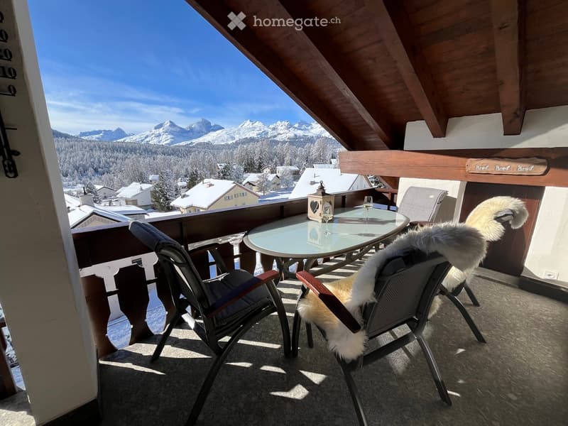 Balcony with a round glass table, two chairs, one with a fur throw, overlooking a snowy mountain landscape.
