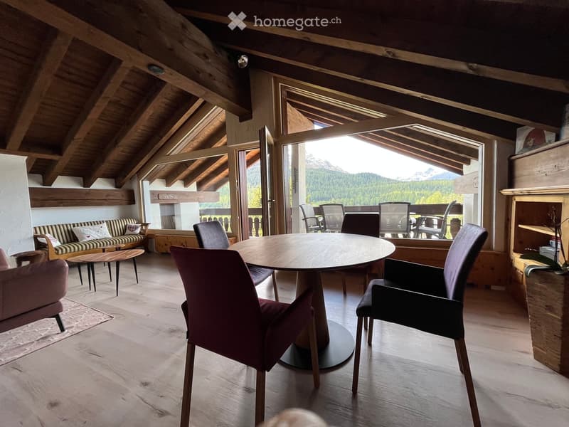 Bright, airy room with wooden beams and exposed ceiling, featuring a round wooden dining table with chairs, a sofa with pillows, and a side table.