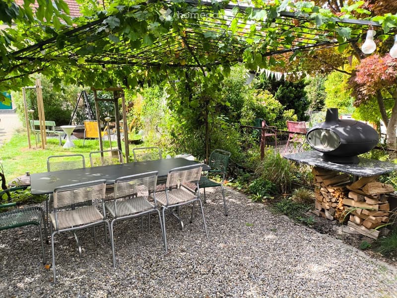 A well-maintained garden with a covered seating area, a dining table with chairs, a stone fireplace, and a stack of firewood.