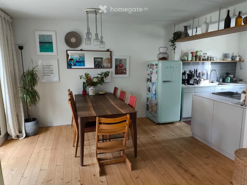 Bright kitchen and dining area with wooden floors, a light blue refrigerator, a wooden dining table with chairs, and open shelving with various items. The space features framed artwork, a potted plant, and a hanging light fixture.