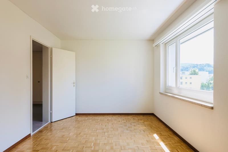 Empty room with light-colored walls, wooden herringbone flooring, and large windows offering a view of the outside. A door is visible on the left side.