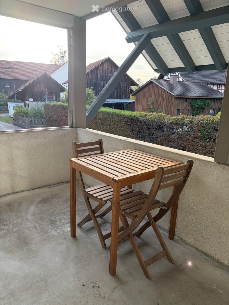 Balcony with a wooden table and two folding chairs, overlooking a residential area with houses and greenery.