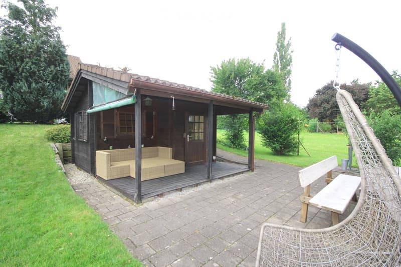 A wooden outdoor structure with a sofa inside, a wooden bench, and a hanging egg chair on a paved patio surrounded by a grassy lawn and trees.