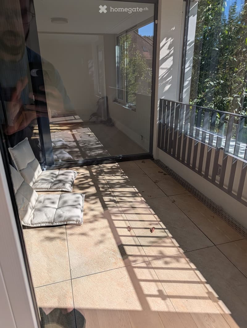 Modern balcony with tiled floor, large windows, and a single white cushioned chair.