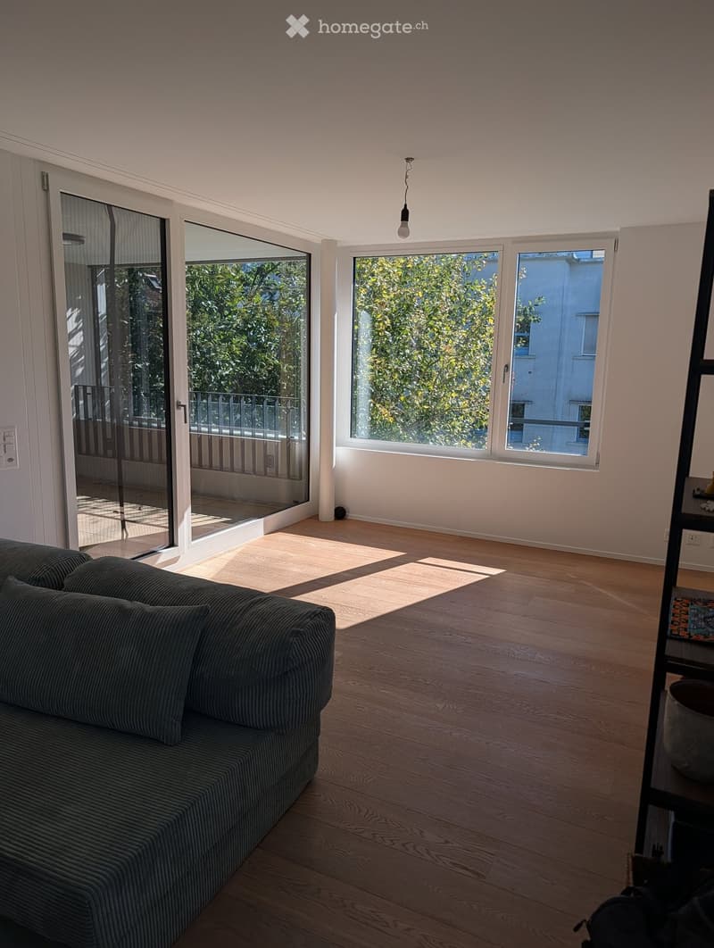 Bright living room with wooden flooring, large windows, and a sliding glass door leading to a balcony. Features a dark grey sofa and a pendant light fixture.