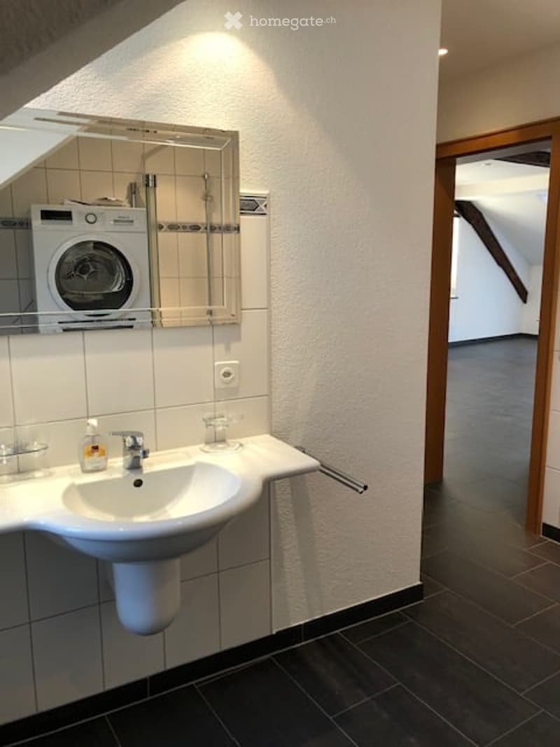 Modern bathroom with white tiled walls, a white sink with a chrome faucet, a mirror above it, and a private washing machine on a shelf. The floor is dark tiled, and the room has a sleek, contemporary design.