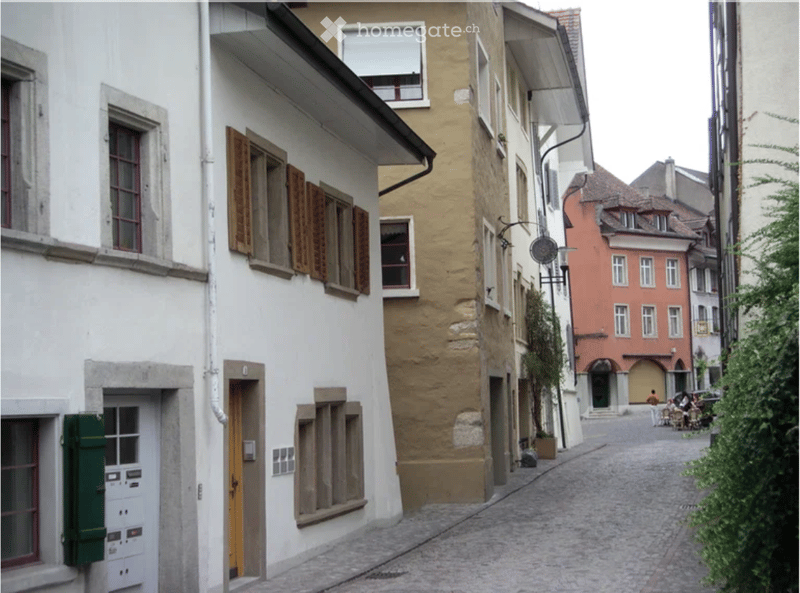 A narrow cobblestone street lined with historic buildings featuring shutters, windows, and doors. The architecture includes a mix of white and beige facades with traditional European design elements.