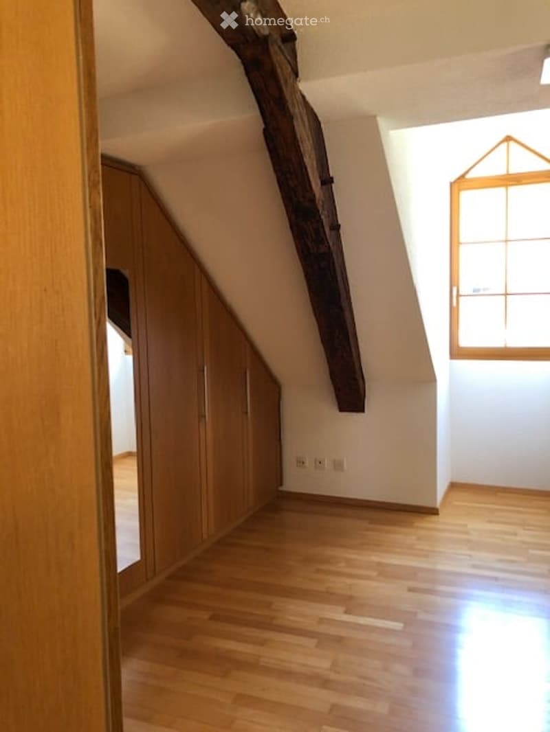 Empty attic room with wooden floor, built-in wooden cabinets, exposed wooden beam, and a single window letting in natural light.