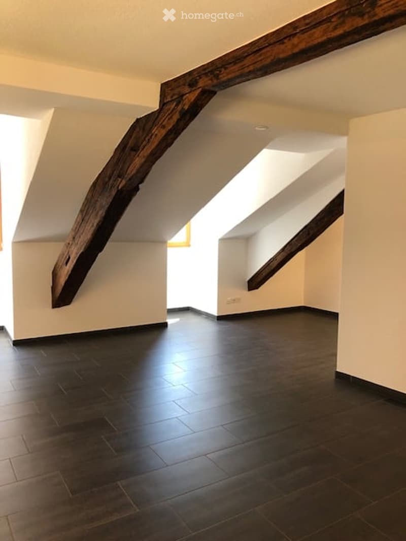 Empty attic space with exposed wooden beams, dark wooden flooring, and white walls. Natural light streams in from a small window at the far end.