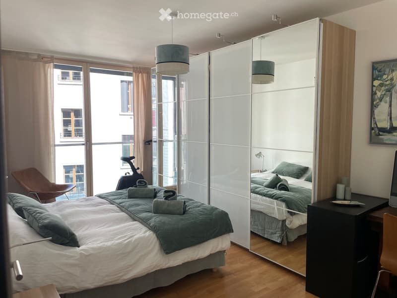 Modern bedroom with a large bed, green bedding, a sliding mirror wardrobe, a wooden chair, and a small table with a lamp.