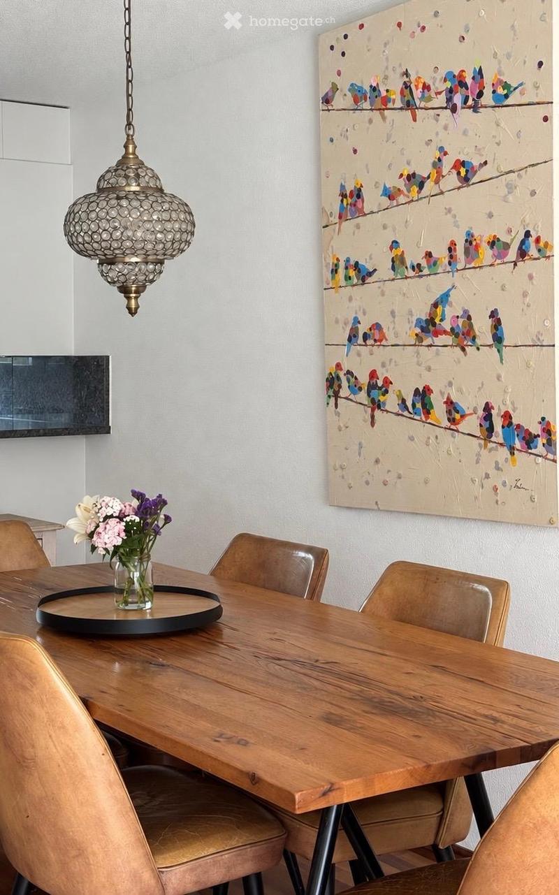 Dining room with a wooden table, four chairs, a vase with flowers, and a colorful bird painting on the wall.
